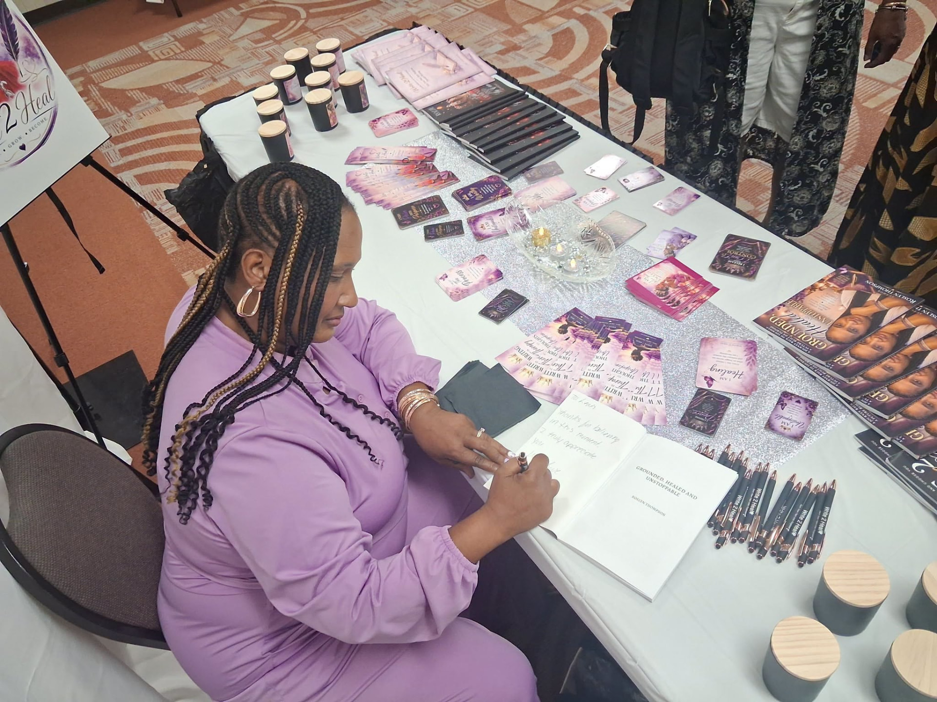 Roslyn Thompson seated at the Write 2 Heal vendor table, signing a copy of Grounded, Healed & Unstoppable beside stacks of journals and purple affirmation cards.