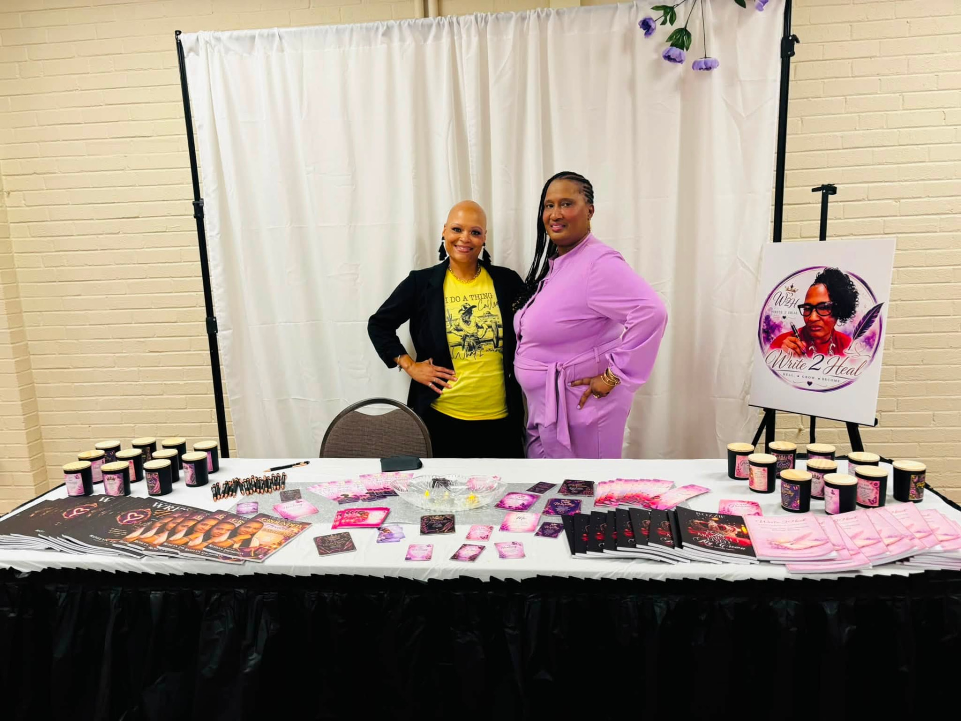 Two guests posing behind the Write 2 Heal table of books, candles, and bookmarks.